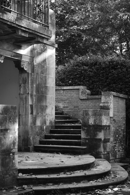 A wide view of Gunnersbury Park showing a sunny outdoor area with large, leafy green trees providing shade and a well-maintained grassy verge. In the background, a series of stone steps and open balus
